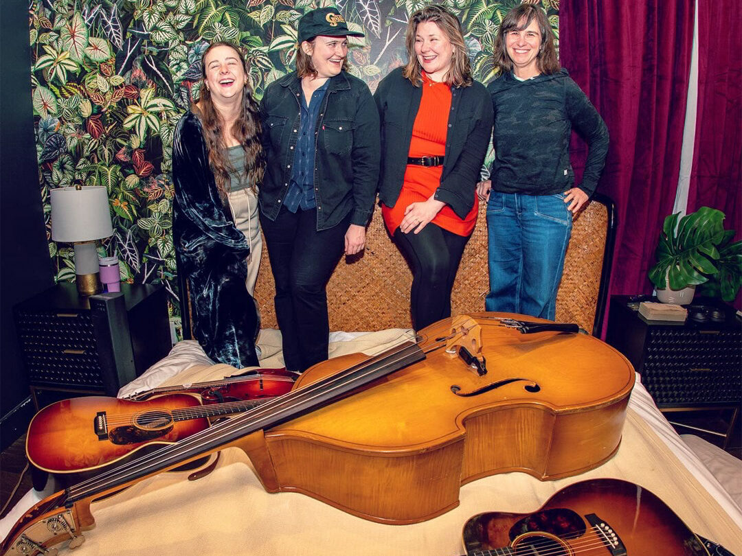 Della Mae bandmembers lined up in the background, string instruments in the foreground